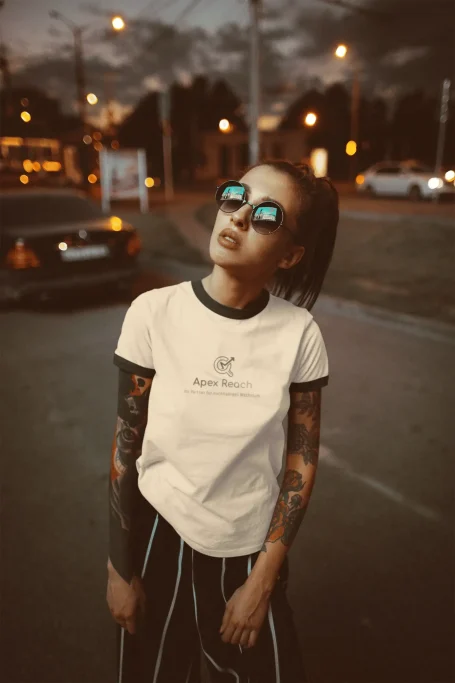 Junge Frau mit Sonnenbrille, Apex Reach T-Shirt und tätowierten Armen vor einer Stadtlandschaft.