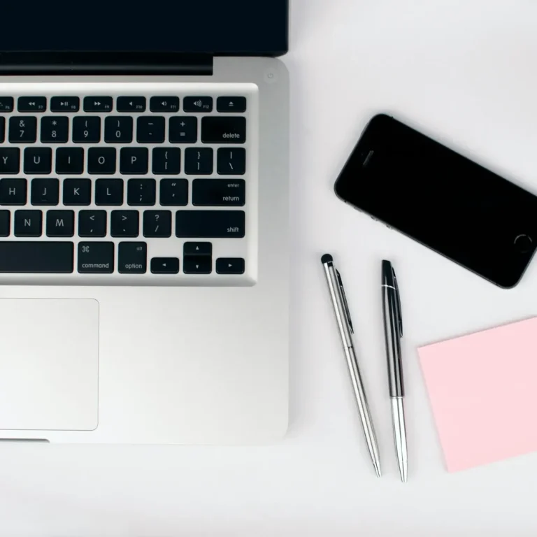 Laptop, schwarze Smartphone, zwei Stifte und ein Post-it-Block auf einem Tisch.